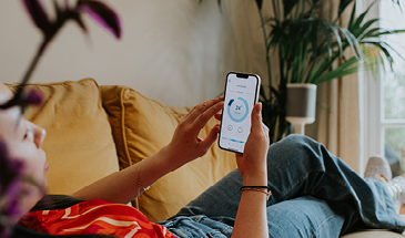 A man lying down on a sofa in his home, he is looking at a heating app on his phone