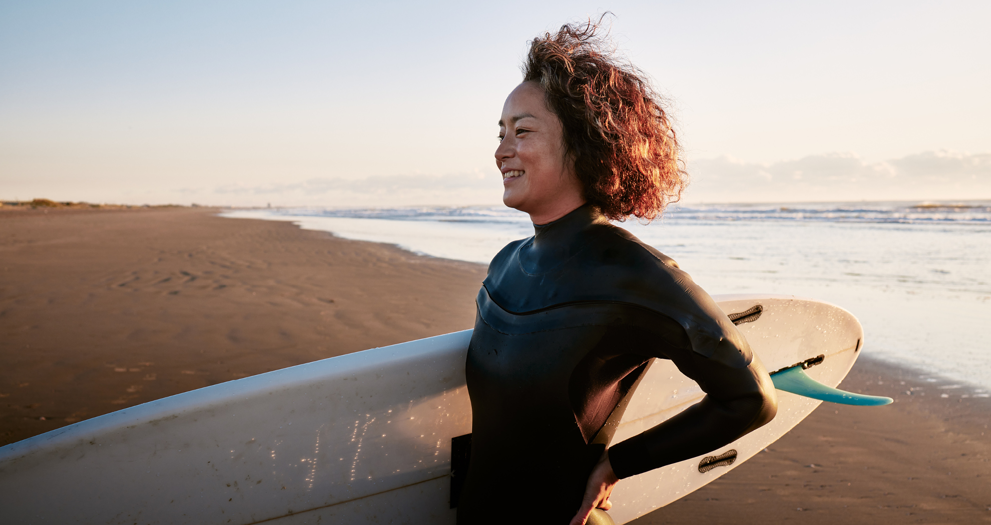 A woman standing on the beach in a wetsuit, holding a surf board
